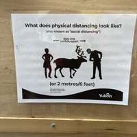 This is a picture of a sign which reads: "What does physical distancing look like? (also known as 'social distancing') Stay one caribou apart (or 2 meters/six feet). A visual representation shows two people on either side of a caribou.