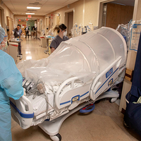 A photo of two healthcare workers moving a hospital bed with a large dome-like covering on top of it.