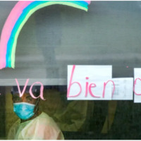 A medical worker looking out a window.  Written in window marker is the phrase "ca va bien aller" with a rainbow.