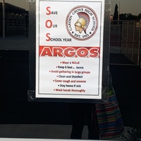 This is a picture taken of a window at a high school. A sign is posted on it which reads: "Save Our School year Argos. Wear a mask, keep six feet distance, avoid gathering in large groups, clean and disinfect, cover cough and sneezes, stay home if sick, wash hands thoroughly." The crest of the school is present on the flyer, which resembles the head of a roman soldier in a classical helmet, and the words "Garden Grove High School Est. 1921" encircling it. 