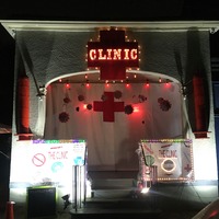 This is a picture of a building that has signs reading "clinic" attached to the front of it. A Red Cross flag is covering the entrance to the building, and small objects resembling COVID-19 particles hang in front of the flag. 