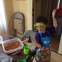 A kid with curly black hair is posing. He has an apron on over his purple shirt and he is wearing green goggles. In front of him is a brown wooden table with a square tupperware with brown water in it.  