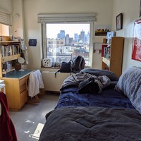 A white dorm room with tile flooring. The room has a window looking out at buildings that continue off into the distance. To the left of the room has a wooden desk that has a bookcase above it that is filled with books. On the right of the room has a bed that is leading out of frame, the bed has a navy bedspread with a grey folded blanket on it. In front of the bed in the corner of the room is another wooden bookcase filled with books. 