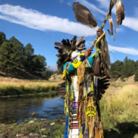 This is a picture of an indigenous man dressed in ceremonial clothing performing a type of traditional ceremony by a creek. 