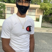 This is a picture taken of a man standing outside in a residential street, wearing a face mask. He is wearing glasses, a dark baseball cap, a white shirt, and a gold watch. 