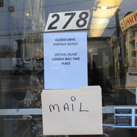 A music store window door with a sign that says "Closed until further notice. Virtual music lessons will take place" and a manilla folder attached underneath with the word mail on it. 