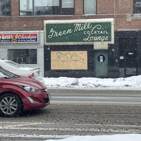 This is a picture taken of a sign for a cocktail lounge from the street on a snowy day. The sign reads "Green Mill Cocktail Lounge".