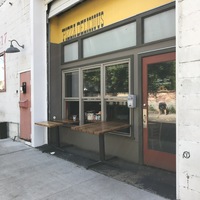 A dark grey and yellow building that says: PIZZA DELICIOUS across the top. The windows on the front of the building have wooden tables hanging out of them.