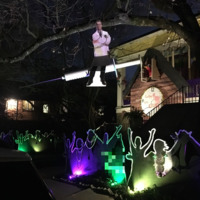 Exterior of home in New Orleans at night, lit with purple, green, and yellow lights.  Cardboard cut outs of people raising hands and dancing are on the ground, and hanging above, suspended from a tree, is a cardboard cut out of a doctor. 