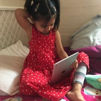 A girl with pigtails in a white and red polka dot dress is sitting on a bed with an iPad sitting in her lap. 