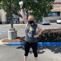 This is a picture of a woman wearing a grey tank top, black pants, glasses, and a face mask. She is standing in a parking lot holding what appears to be a vaccination card above her head. 