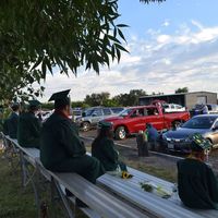 High school graduates sit socially distanced on bleachers outdoors while all attendees watch from their vehicles.