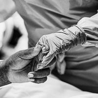 Black-white image of a dark-skinned hand holding the gloved hand of a person who is in full surgical gear. 
