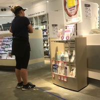 A woman in a pharmacy dressed in all black workout gear wearing a visor and is waiting in line. On the floor is a 6 feet piece of blue tape, measuring social distancing. 