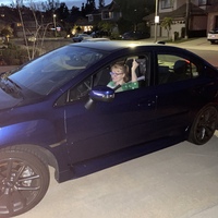 This is a picture of a woman in a green shirt and glasses smiling while raising her pinky finger in the air out of her car window in a driveway. The background of the photo is a residential neighborhood at night. 