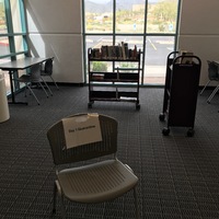 The corner of a library with windows. There are two book carts with books. There is a chair with a paper sign on it that says: Day 1 Quarantine. 