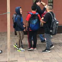 Three kids wearing backpacks are standing in front of a table outside. The kids are talking to one another. Behind the table is a woman who is putting paper lunch bags onto the table. 