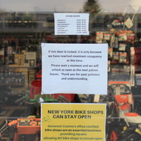 Three signs on a window.
Top sign is store hours.
Middle sign says "If this door is locked, it is only because we have reached maximum occupancy at this time. Please wait a moment and we will unlock as soon as the next patron leaves. Thank you for your understanding and patience." 
Bottom sign says "New York bike shops can stay open. Governor Cuomo's office clarified bike shops are an essential business-providing repairs-allowing NY bike shops to remain open during the Workforce Reduction mandate under Executive Order 202.6."