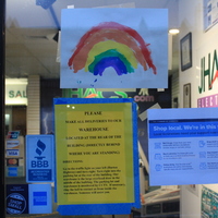 A window with a couple of signs taped to it. There is a sign to shop local on the right, directions to make deliveries to the facilities warehouse on the left, and a rainbow painting that sits above both signs. Through the window, the company JHACS Electric can be seen.