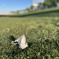 This is a picture of a face mask that has been disposed of in a grassy area. The rest of the picture is out of focus, but a building and trees can be barely seen blurred in the background of the shot. 