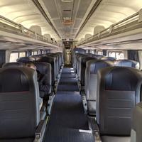 Inside of an Amtrak cabin that only has one person sitting in a seat off to the left. The rest of the cabin is empty. The isle has blue carpet to match the blue headrests of the seats. 