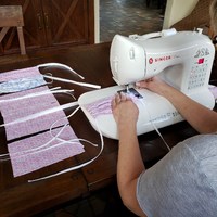 A person is sitting at a table using a white sewing machine sewing face masks. On the table off the left is scrap material for more face masks. 
