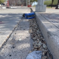 This is a picture of a face mask that has been discarded in a gutter on a street. Buildings can be seen in the blurry background. 