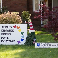 Photo of someone's front yard featuring two gnomes and a sign that reads "April's distance brings May's existence". 