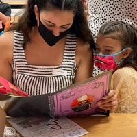 This is a picture of a woman reading from a pink notebook to a younger girl. Both are wearing face masks. 