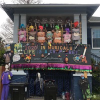 This is a picture taken of a person's house which is decorated with various items related to the COVID-19 Pandemic. A line of poop shaped emojis with smiley faces hang from the roof, under which sits a line of dummies with skulls for heads each labeled under a different spelling of the name "Karen", with a sign underneath stating that each dummy did not wear their mask. A banner underneath these objects reads "2020 in musicals: 'Life is a Crap-aret'". The bottom of the banner encourages the reader to follow social distance protocols. Several mannequins dressed in medial gear or face masks flank the banner. 