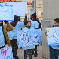 This is a picture of face masked children holding signs that protest the Neskantaga water crisis. One sign reads "water is sacred." 